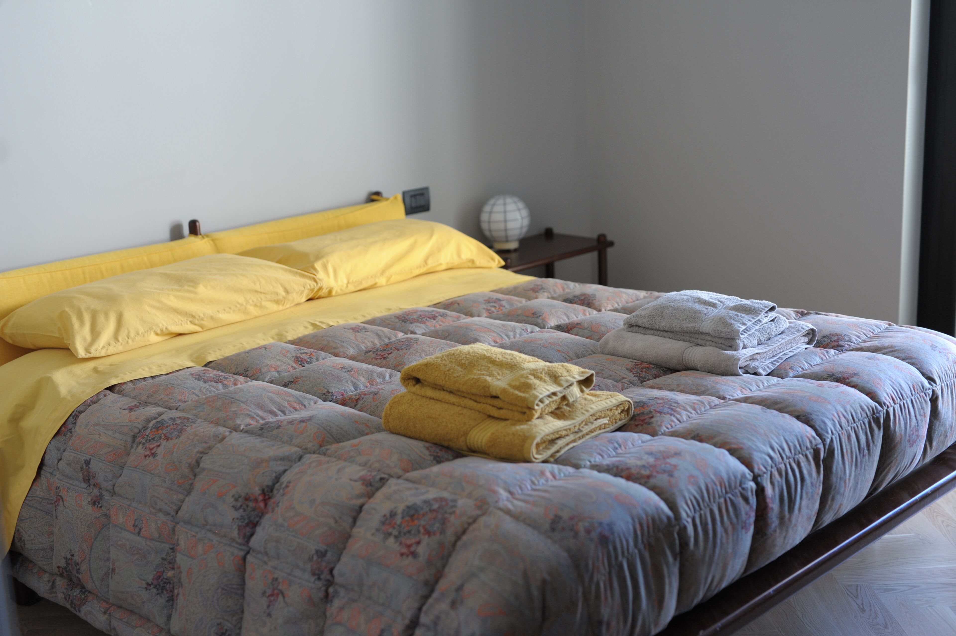 A neatly made bed with a floral-patterned duvet, a yellow blanket, and folded towels.