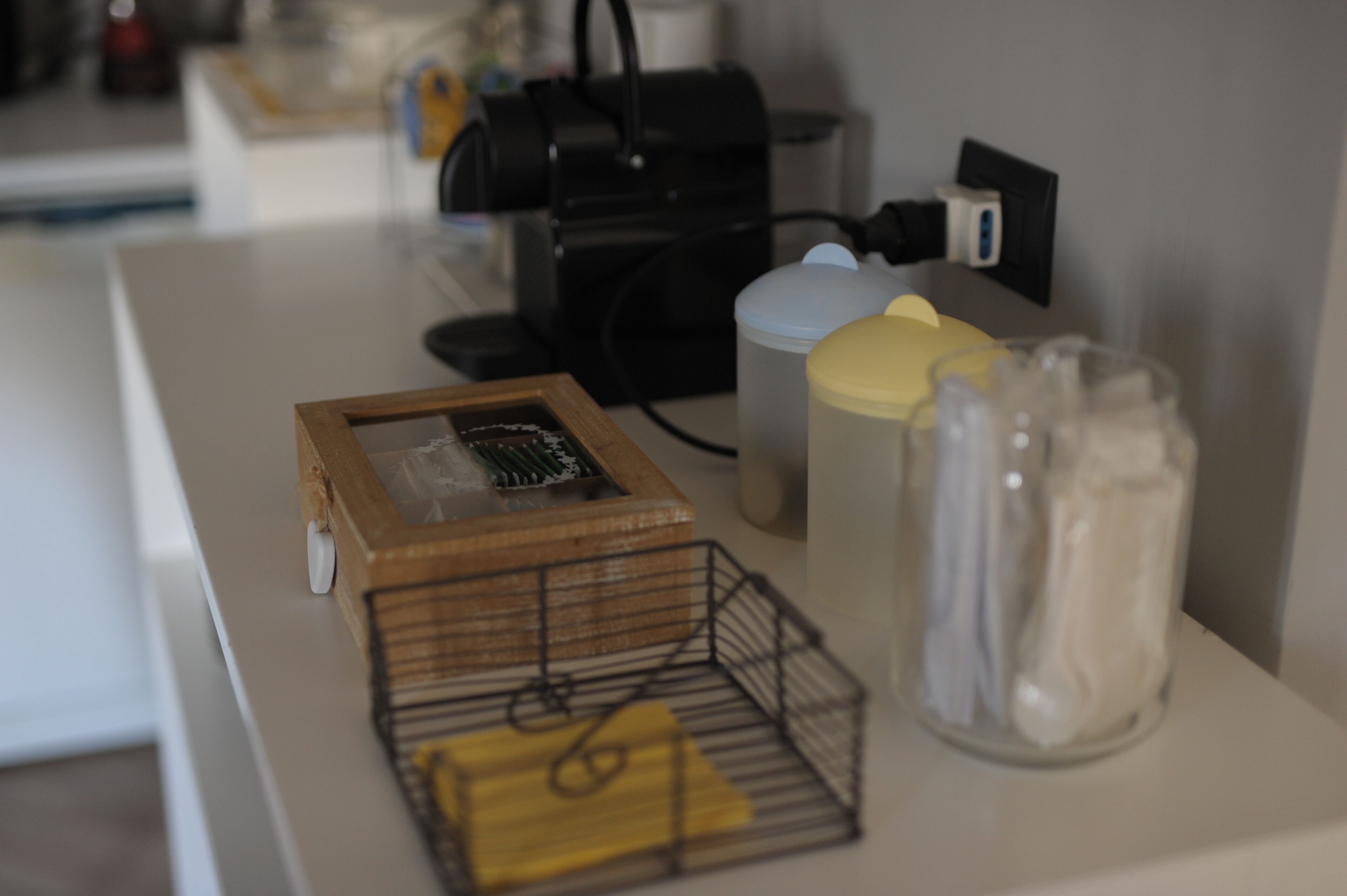 Uma bancada de cozinha com uma cafeteira, um recipiente com tampa, uma pequena caixa de madeira e um pote de vidro com tampa de metal.
