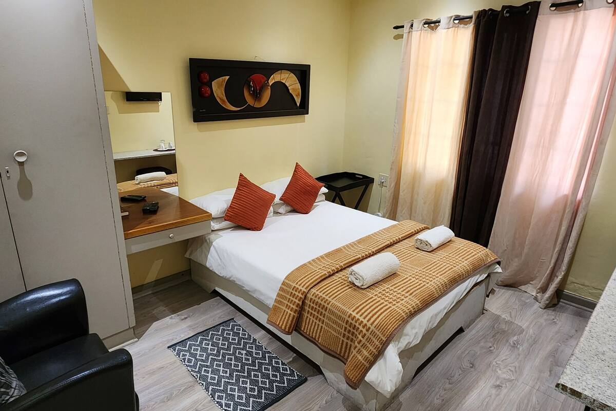 A hotel room with a bed, a desk, a chair, a small table with cups, and a potted plant.