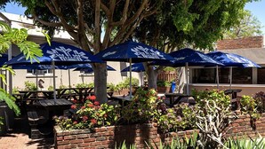 A patio area with outdoor seating under blue umbrellas, surrounded by lush greenery and a large tree. - Pofadder Hotel (Pofadder)