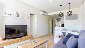 A modern living room with a flat-screen TV, a wooden coffee table, a grey sofa, and a dining area with a white table and chairs.
