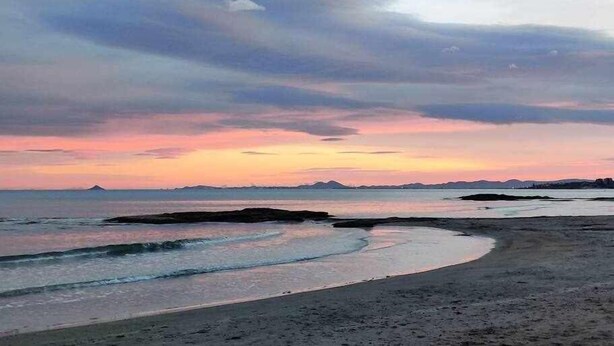 A beach with a curved shoreline and distant mountains under a colorful sky.
