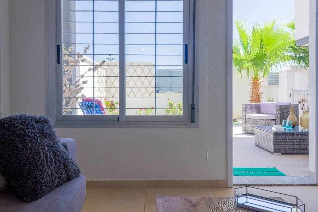Ein Zimmer mit großem Fenster, einer Couch und Blick auf eine Außenpatio-Terrasse mit Möbeln und einer Palme.