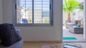 A room with a large window, a sofa, and a view of an outdoor patio area with furniture and a palm tree.