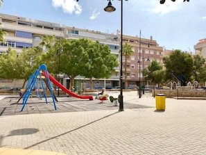 Un parque infantil con un tobogán rojo y una estructura de escalada azul, rodeado de árboles y edificios.