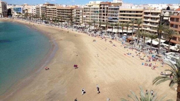 Une plage bordée d’immeubles à plusieurs étages, de palmiers et sous un ciel dégagé.
