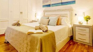 Une chambre à coucher avec un grand lit, une table de chevet avec une lampe, une commode et un tableau représentant une scène de plage.