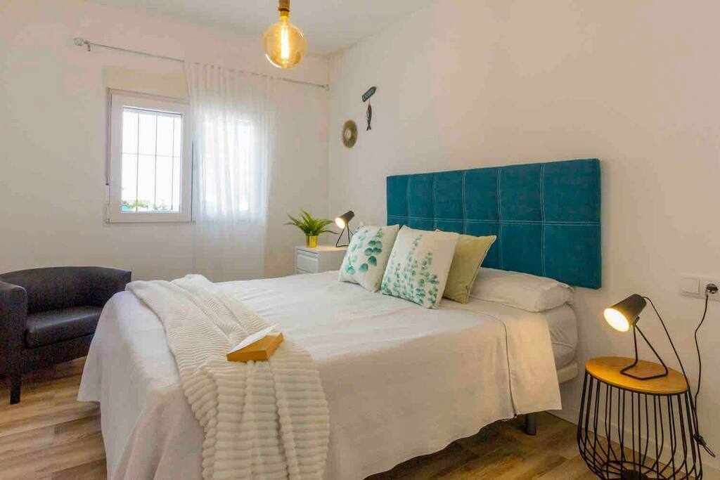A bedroom with a bed, a chair, a bedside table with a lamp, and a window with curtains.
