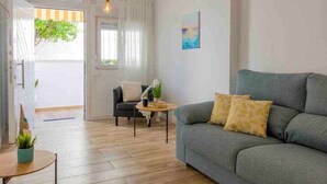 Ein Wohnzimmer mit grauem Sofa, einem kleinen runden Tisch und einem Stuhl. Es gibt ein Fenster mit Vorhängen, eine Tür und eine Pflanze auf dem Tisch.