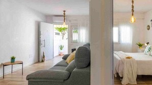 Ein modernes Wohnzimmer mit einem grauen Sofa, einem kleinen Tisch mit Pflanze und einem Bett mit weißer Decke.