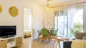 A modern living room with a flat-screen TV, a dining area with chairs, a sofa, and a large window with curtains.