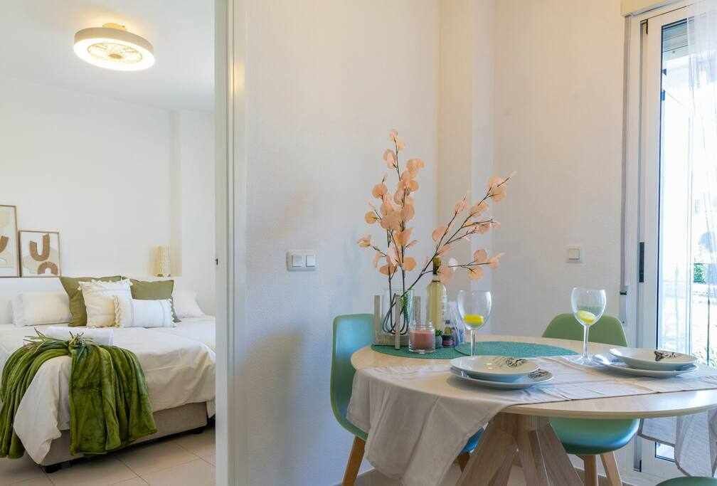 A room with a bed, a dining table with chairs, and a vase with flowers.