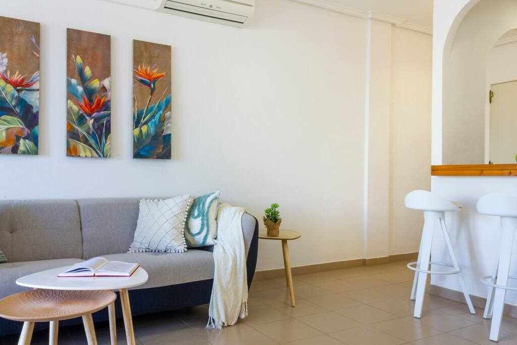 A modern living room with a grey sofa, a small round table with a book, and two white bar stools.