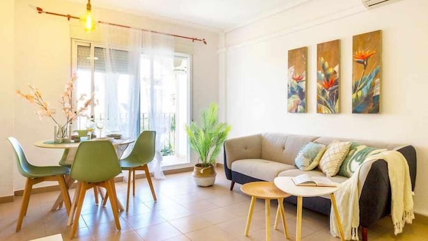 A modern living room with a sofa, a dining table with chairs, and a small coffee table. There is a potted plant and artwork on the wall.