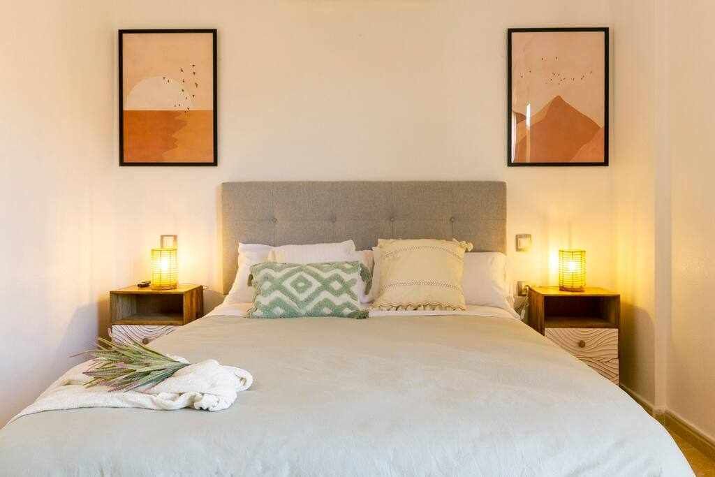 A neatly made bed with a grey headboard, two bedside tables with lamps, and two framed artworks on the wall.