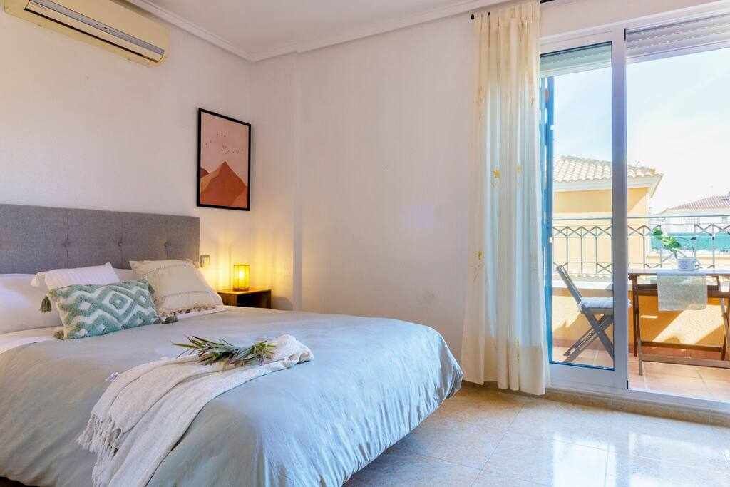 Een slaapkamer met een bed, een nachtkastje, een lijst met een afbeelding en een balkon met een tafel en stoelen.