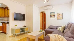 Una sala de estar con un sofá, una mesa de centro y un televisor montado en la pared. Se ve una cocina a través de un arco.