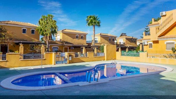 Una piscina rodeada de edificios amarillos con balcones, palmeras y un cielo azul despejado.