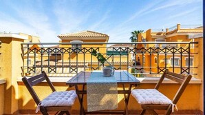 Un balcón con dos sillas y una mesa de madera, baranda de metal y vista de edificios cercanos.