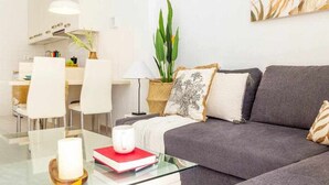 Ein modernes Wohnzimmer mit einem grauen Sofa, einem Glastisch und einem Essbereich mit weißen Stühlen.