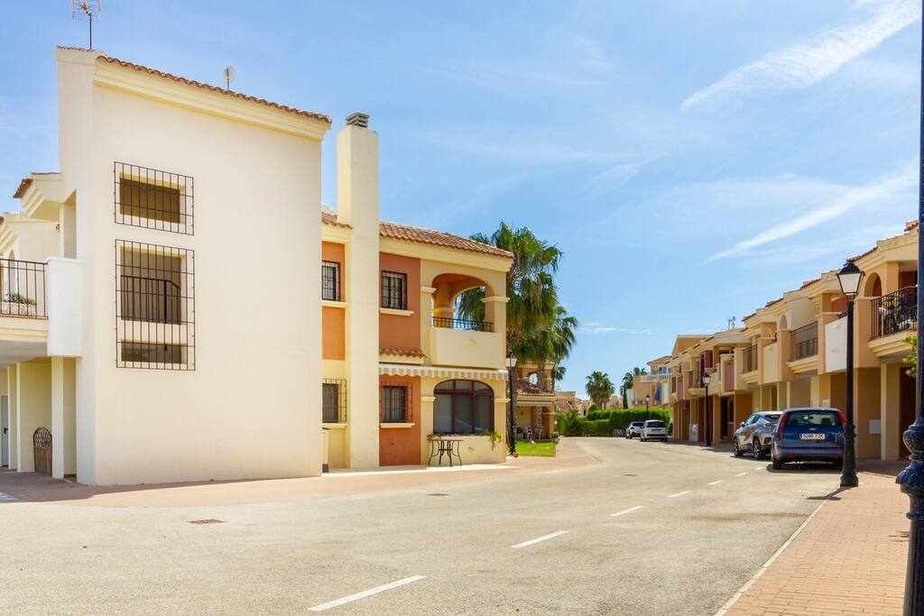 A residential street with a two-story building featuring balconies, a clear sky, and parked cars.