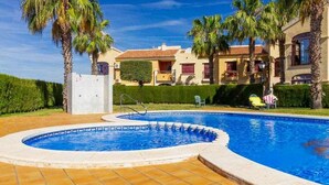 A swimming pool with a tiled deck, surrounded by palm trees and a building with a tiled roof. - Lovely Groundfloor with swimming pool (Torrevieja)
