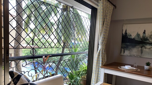 Una habitación con un sofá blanco, una reja metálica en la ventana, una mesa de madera con un bol y un tarro, y un cuadro en la pared.