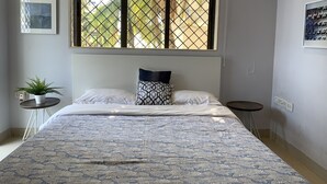 Un dormitorio con cama, ventana con vistas, una mesita con una planta y una lámpara fijada a la pared.