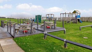 Un parque infantil con estructuras de madera, un área cercada y vista al agua de fondo.