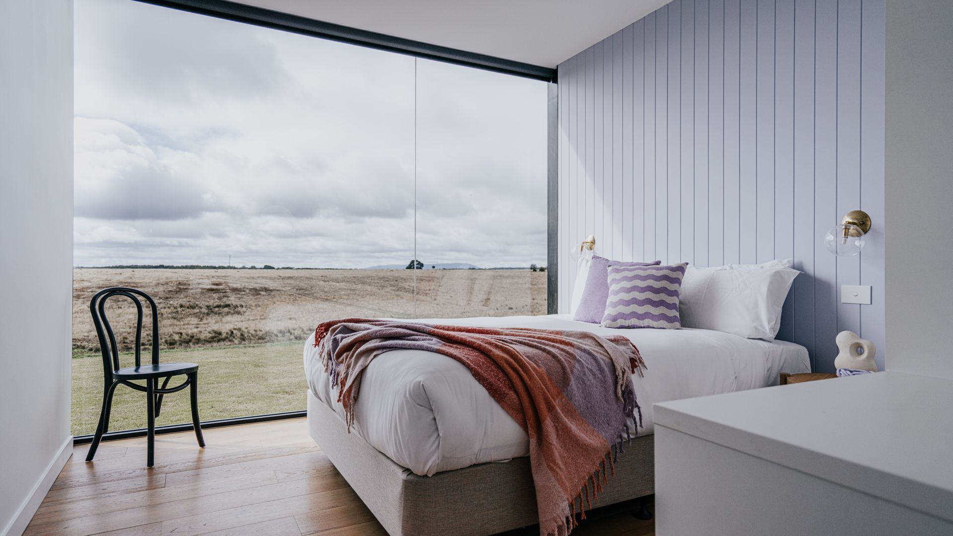 A bedroom with a large window, a bed with a blanket, a chair, and a bedside table.