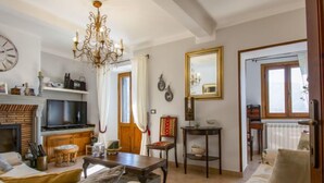 A cozy living room with a fireplace, television, and a chandelier. There is a dining area with a table and chairs, and a window with curtains. - Vacation home Kenzo by Interhome (Bagni di Lucca)