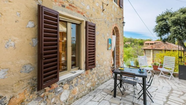 Un bâtiment en pierre doté d’un patio comprenant une table et des chaises, une fenêtre avec des volets et une vue sur le paysage environnant.