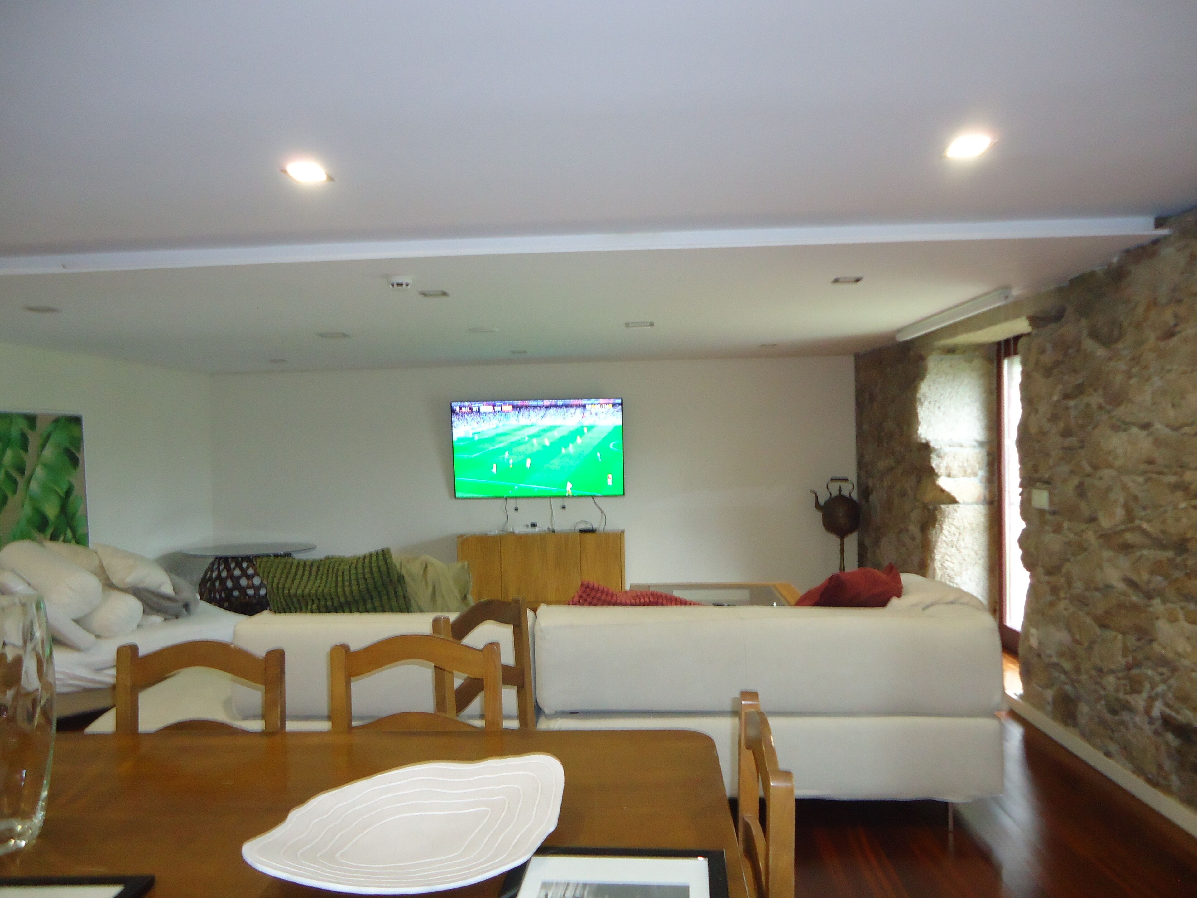 A living room with a TV mounted on the wall, a wooden dining table, and a stone wall.