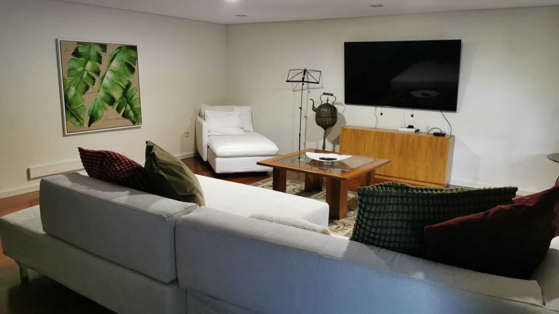 A modern living room with a white sofa, a coffee table, a white armchair, a flat-screen TV mounted on the wall, and a decorative plant.