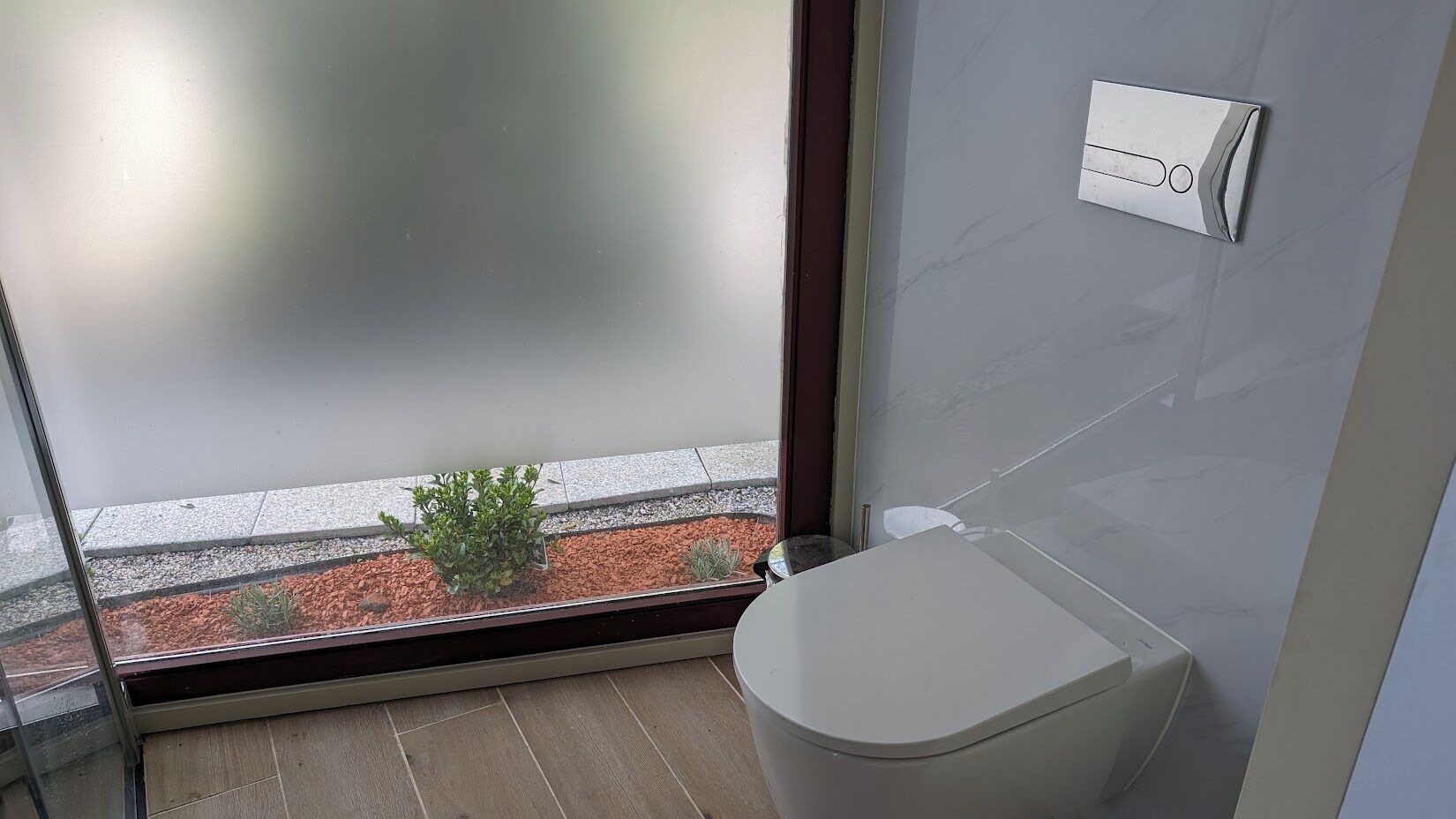 Un baño moderno con inodoro, una planta pequeña y ventana con vista a un jardín.