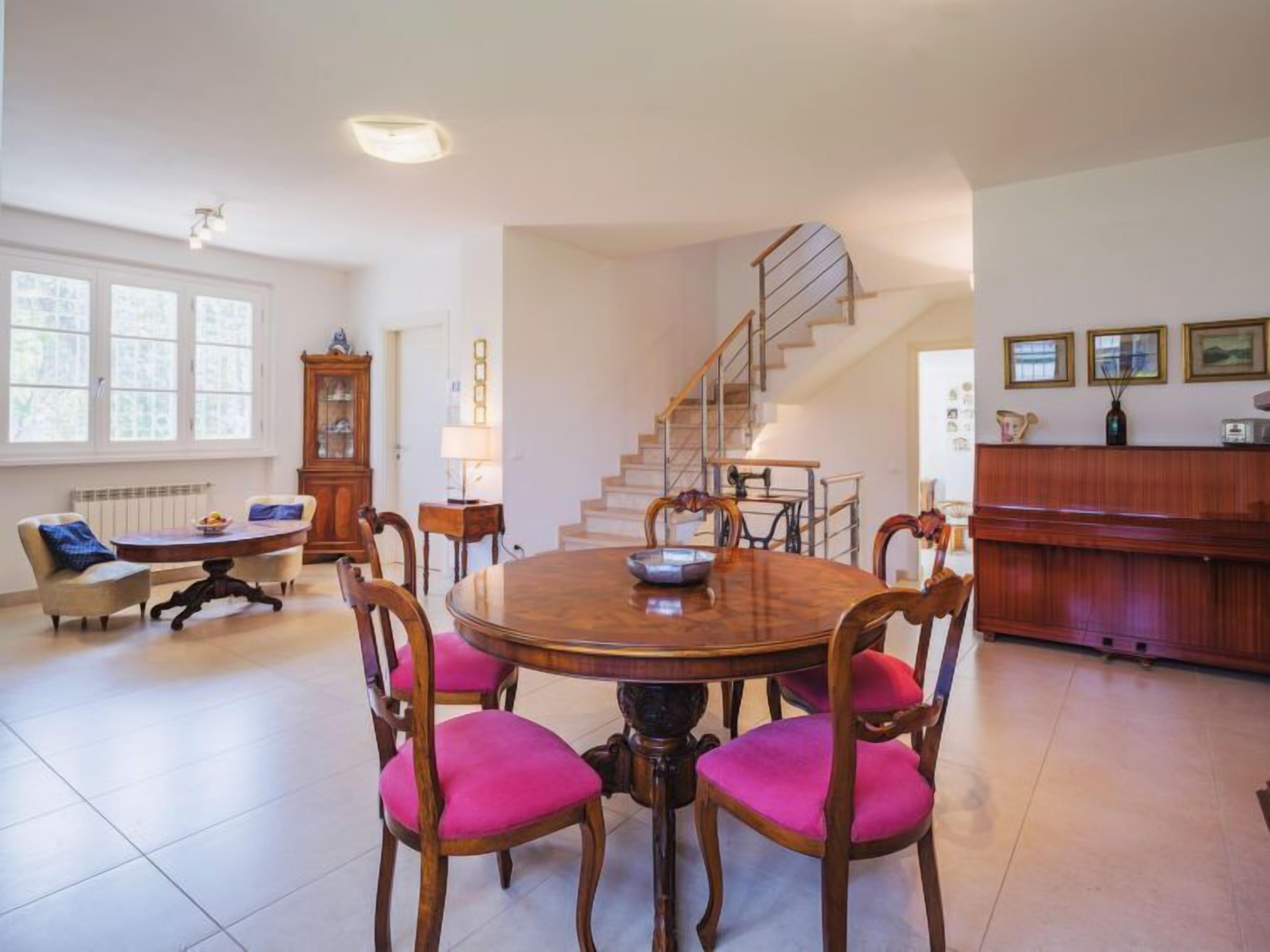 A dining area with a round wooden table, six chairs with pink cushions, a wooden cabinet, and a staircase leading to another level.