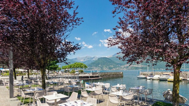 An outdoor café with tables and chairs, a body of water, boats, and mountains in the background. - La Perla by Interhome (Colico)