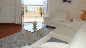 Een woonkamer met een witte bank, een glazen salontafel en uitzicht op een balkon met een hekwerk.