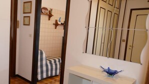 Un couloir doté d’un sol à carreaux, une armoire blanche sur laquelle repose un objet bleu, un miroir à motifs rayés et une porte ornée d’un cadre décoratif.