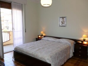 Ein Schlafzimmer mit einem Bett, zwei Nachttischen mit Lampen, einem gerahmten Bild an der Wand und einer Schiebeglastür zu einem Außenbereich.