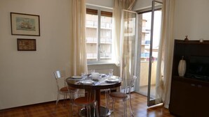 Un coin repas avec une table ronde dressée pour quatre personnes, des chaises transparentes, un plancher en bois, une porte-fenêtre coulissante donnant sur un balcon et un téléviseur posé sur un meuble.