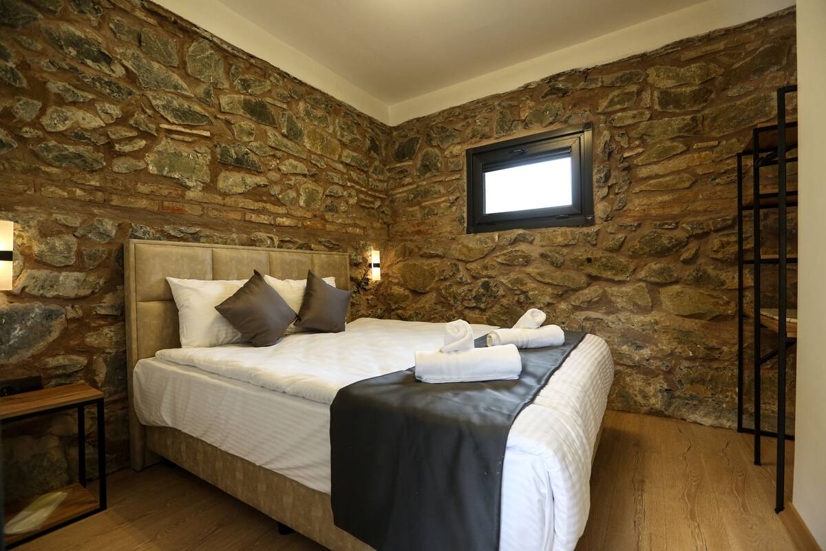 A bedroom with a stone wall, a bed with white bedding, a black throw, and a wooden nightstand.