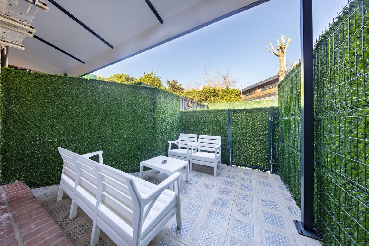 A covered patio area with white outdoor furniture, a patterned floor, and a green hedge wall.