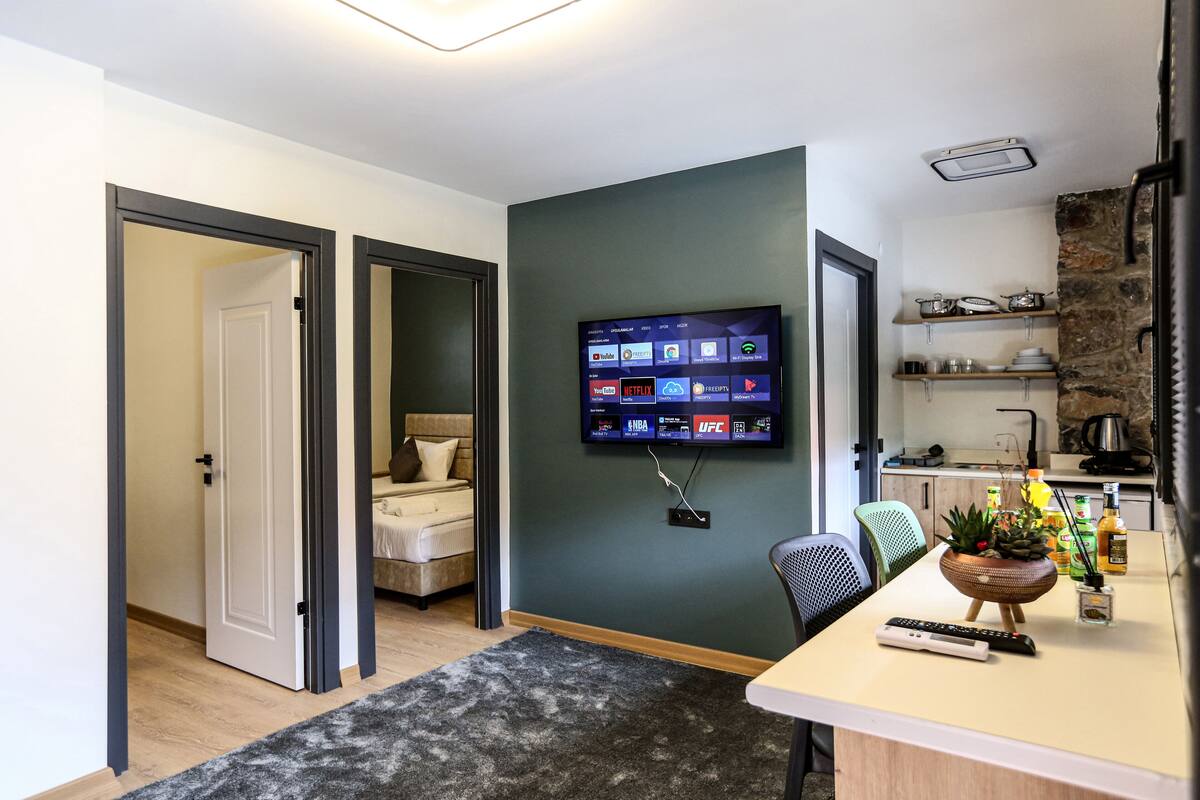 A modern living room with a flat-screen TV mounted on the wall, a sofa, a coffee table, and a kitchen area with shelves and a refrigerator.