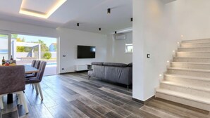 Uma sala de estar moderna com um sofá cinzento, uma TV de tela plana e uma área de jantar com uma mesa e cadeiras.