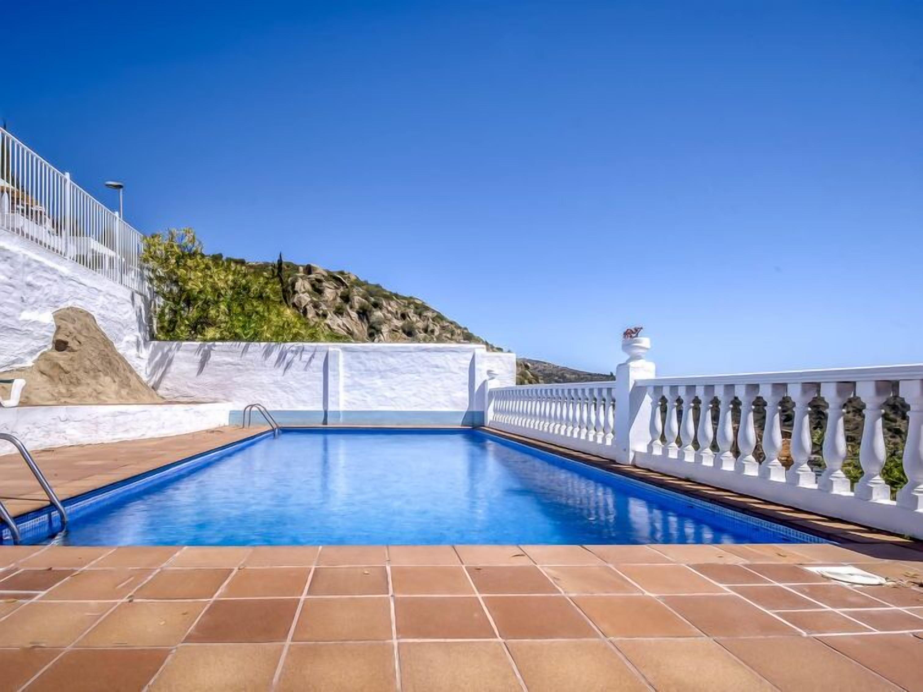 Una piscina con ringhiera bianca, solarium piastrellato e vista su un pendio roccioso.