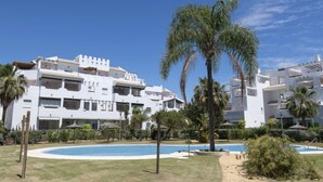 Un immeuble blanc à plusieurs étages, doté de balcons, d’une piscine et de palmiers.