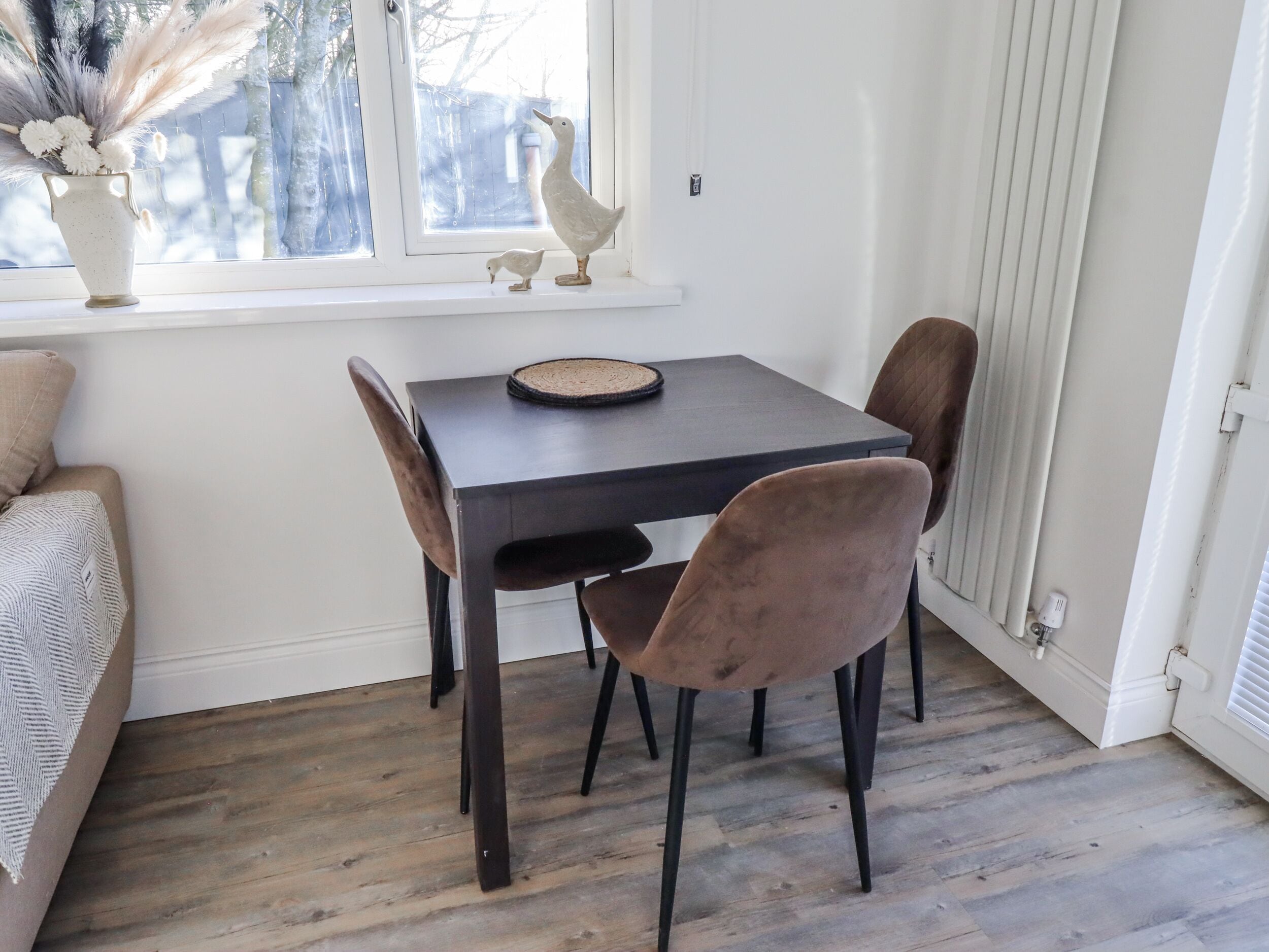 Een zithoek met een tafel en stoelen, een vaas met gedroogde bloemen, en een raam met uitzicht.