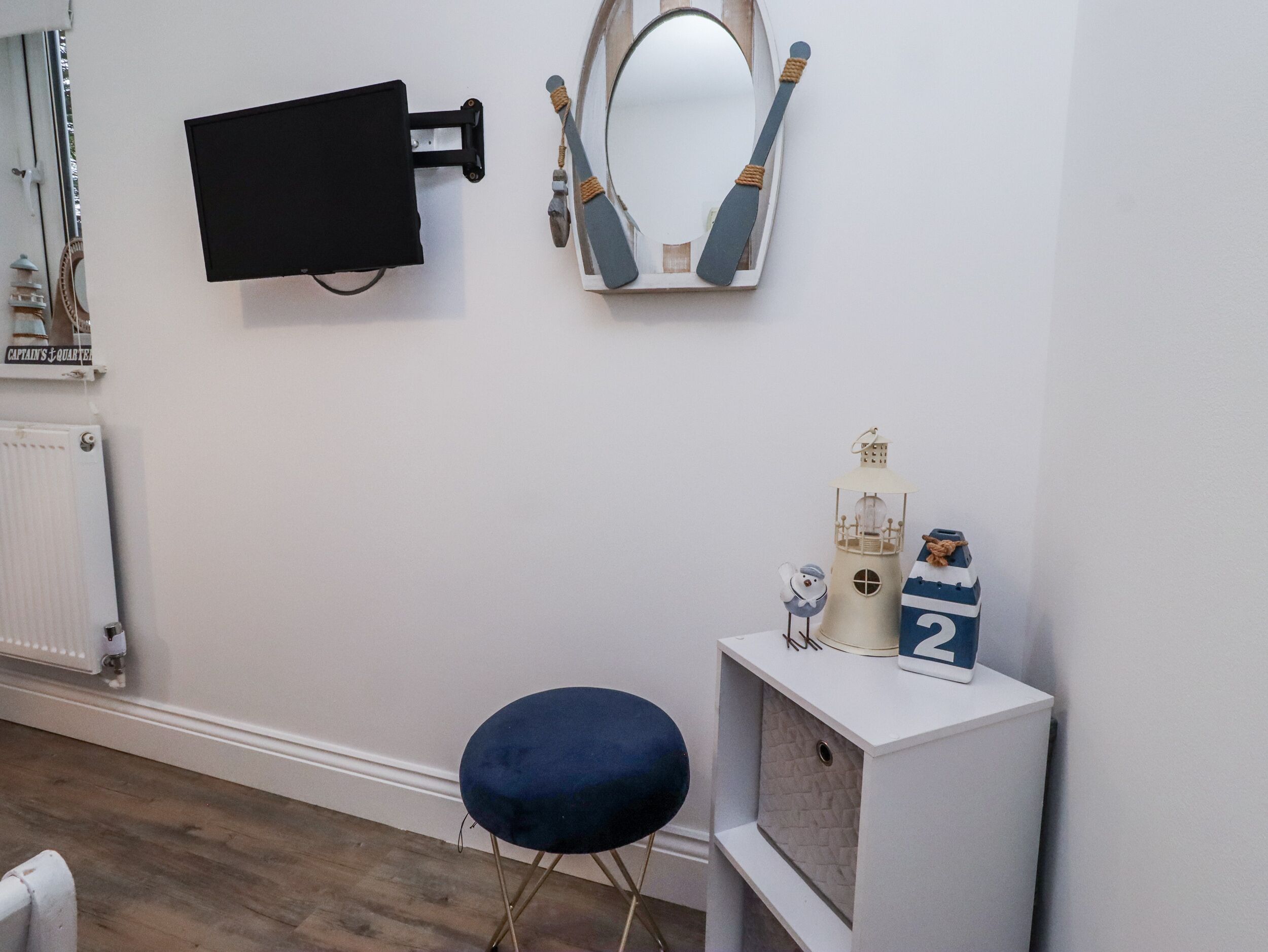 A room with a white radiator, a black TV mounted on the wall, a mirror with a wooden frame, a blue cushioned stool, and a small white shelf with decorative items.