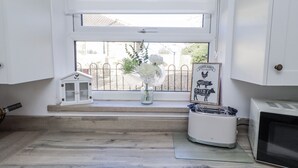 Una pequeña cocina con ventana, un refrigerador blanco, un microondas, un pequeño objeto con forma de casa y un cuadro en el alféizar.